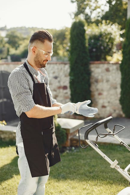 A man with neatly styled hair, glasses, and a trimmed beard is outdoors in a well-maintained garden during daytime. He is wearing a checkered shirt, a black gardening apron, and white gloves, appearin