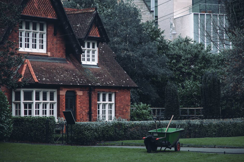 A traditional red brick house with a steep, tiled roof and white-framed windows, situated within a well-maintained garden. The garden features a neatly trimmed hedge bordering a grassy lawn that is be